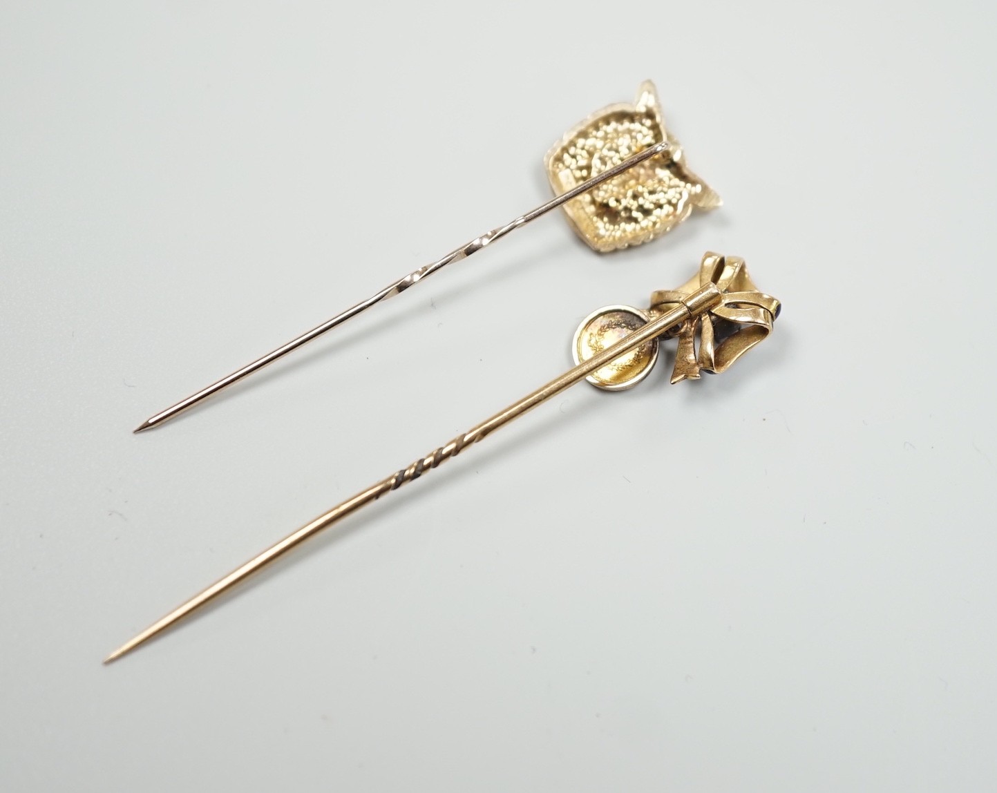 An early 20th century yellow metal enamel and diamond set ribbon bow stick pin, hung with a miniature medallion, 63mm and a later 9ct gold stick pin, gross 5.2 grams.